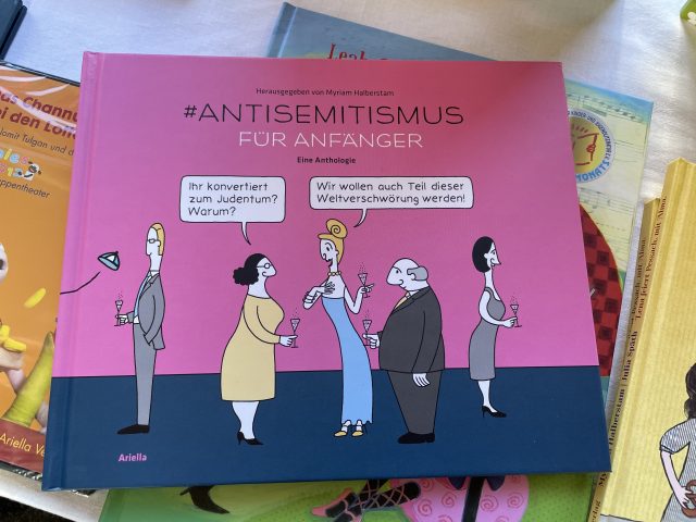 Das Buch "#Antisemitismus" liegend auf anderen Büchern. Hauptsächlich ist das Cover rosa mit dem Titel und 5 Personen auf blauem Grund mit zwei Sprechblasen. 