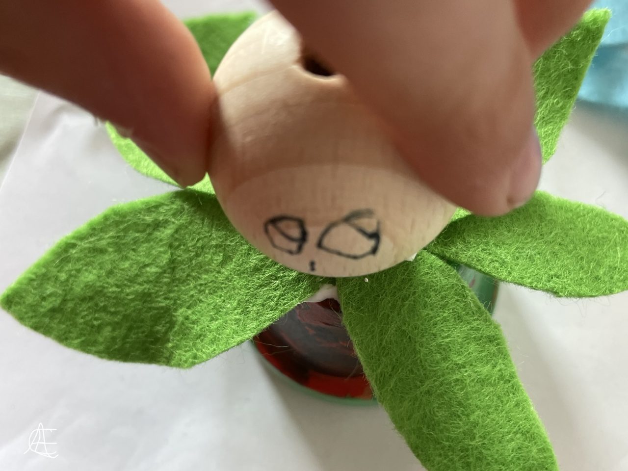 Kinderfinger setzen eine mit Gesicht versehene Holzkugel auf Filzblätter auf der Unterseite eines bunten Tontöpfchens