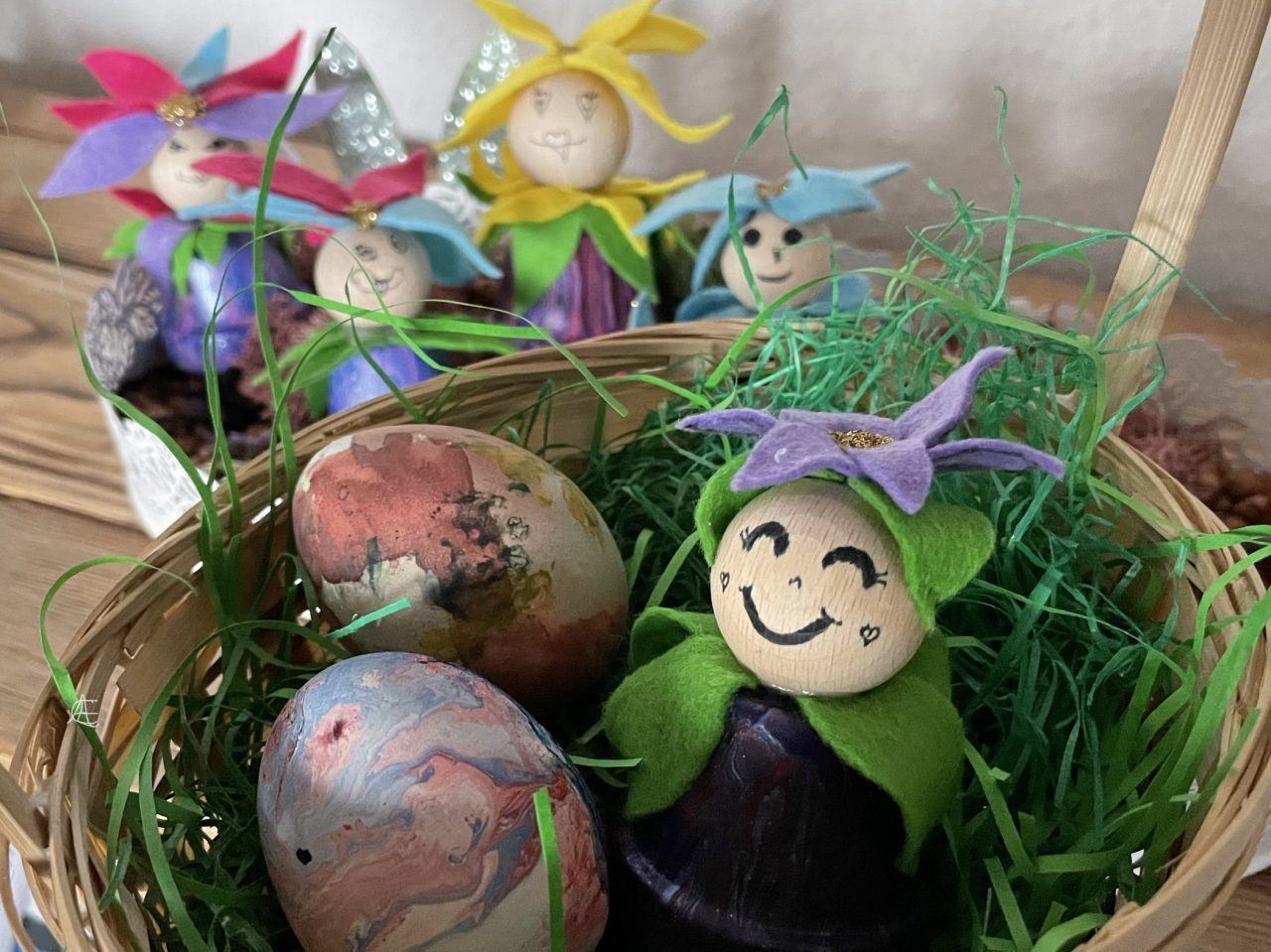 Blümling (Bastelei aus Tontöpfchen, welche an eine Blume erinnert) in einem Körbchen mit Ostergrün und zwei Eiern. Im Hintergrund stehen weitere Blümlinge. 