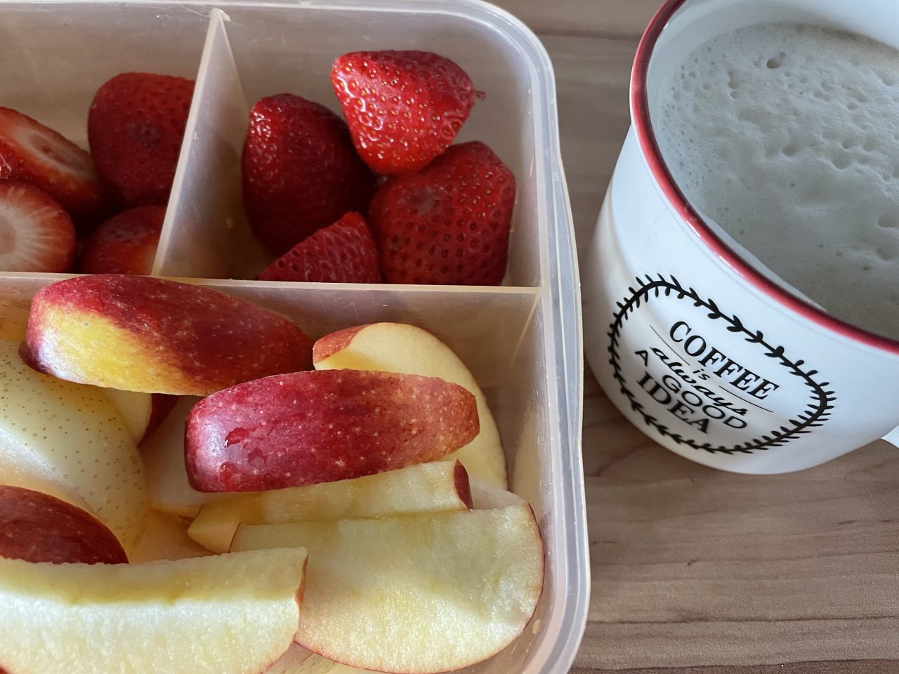 Brotdose mit Erdbeeren und Äpfelschnitzen gefüllt neben einer gefüllten Kaffeetasse