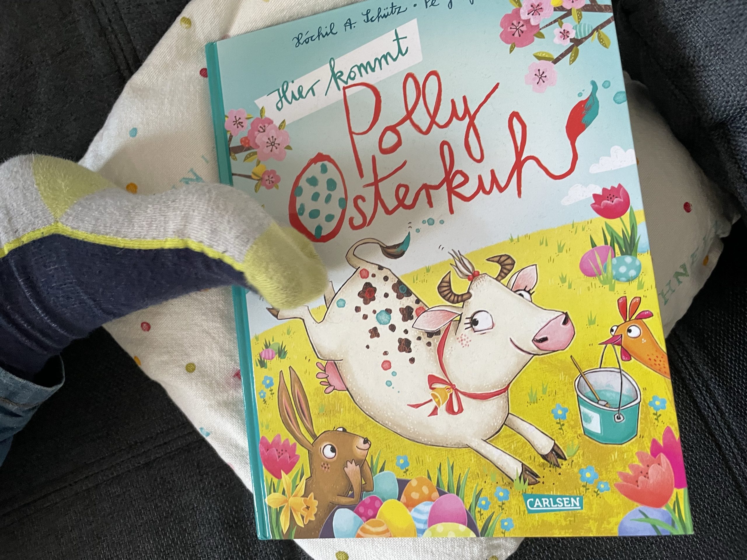 Kinderbuch "Polly Osterkuh" auf dem Sofa mit Kinderfuß