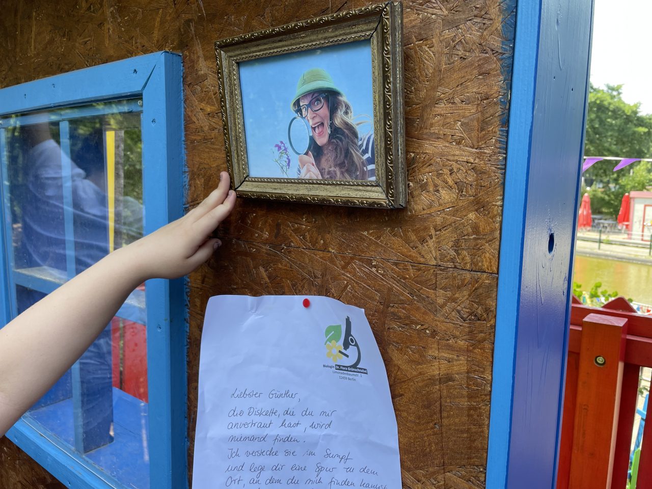 ein Bild und ein Zettel an einer Holzwand befestigt - eine Hand zeigt darauf und im Fenster spiegelt sich etwas weißes