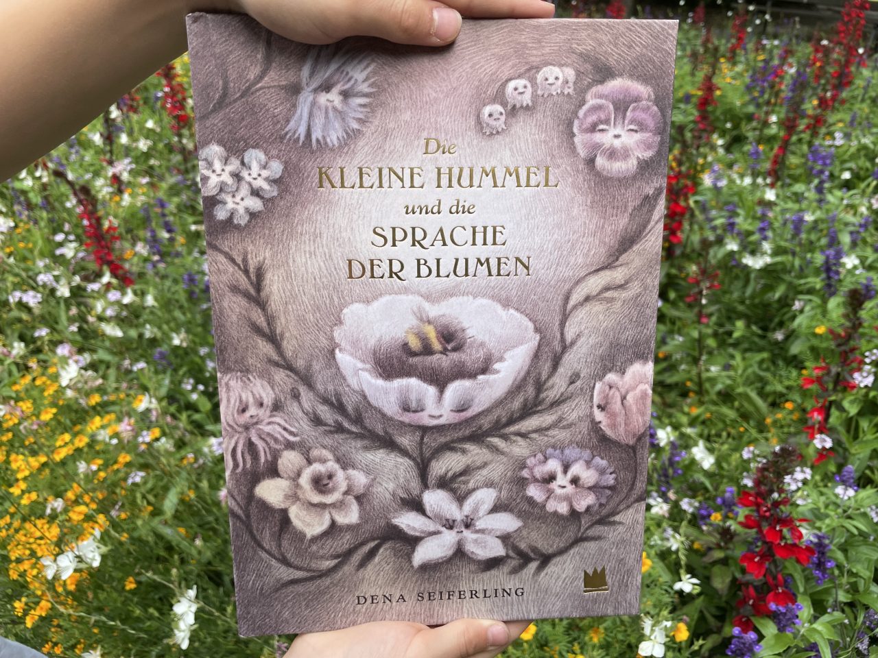 das Kinderbuch "Die kleine Hummel und die Sprache der Blumen" gehalten von zwei Kinderhänden vor einer blühenden Blumenwiese