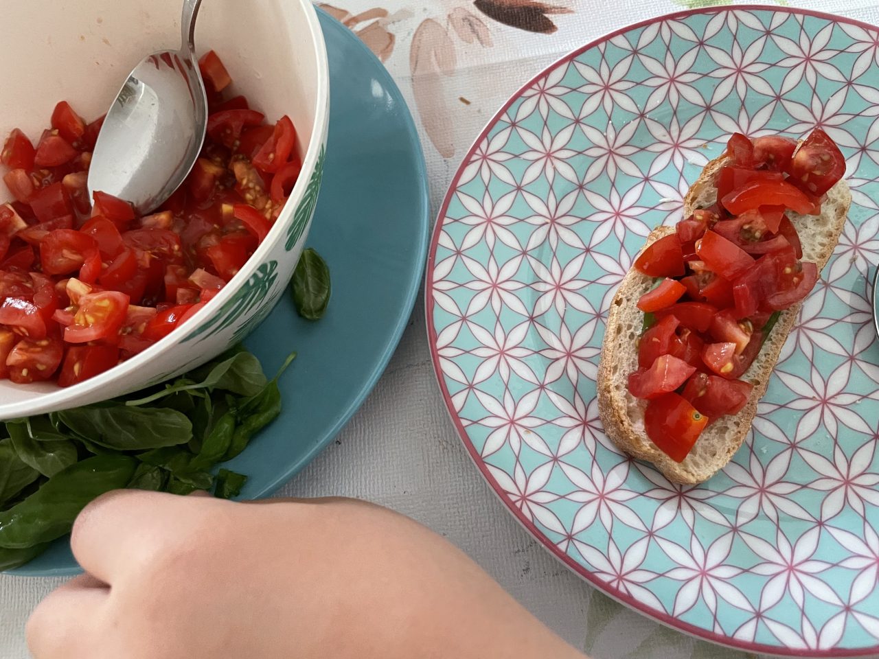 klein geschnittene Tomaten in einer Schüssel auf einem Teller mit Basilikum, dazu ein Teller mit Tomatenbrot und eine Kinderhand mit Löffel