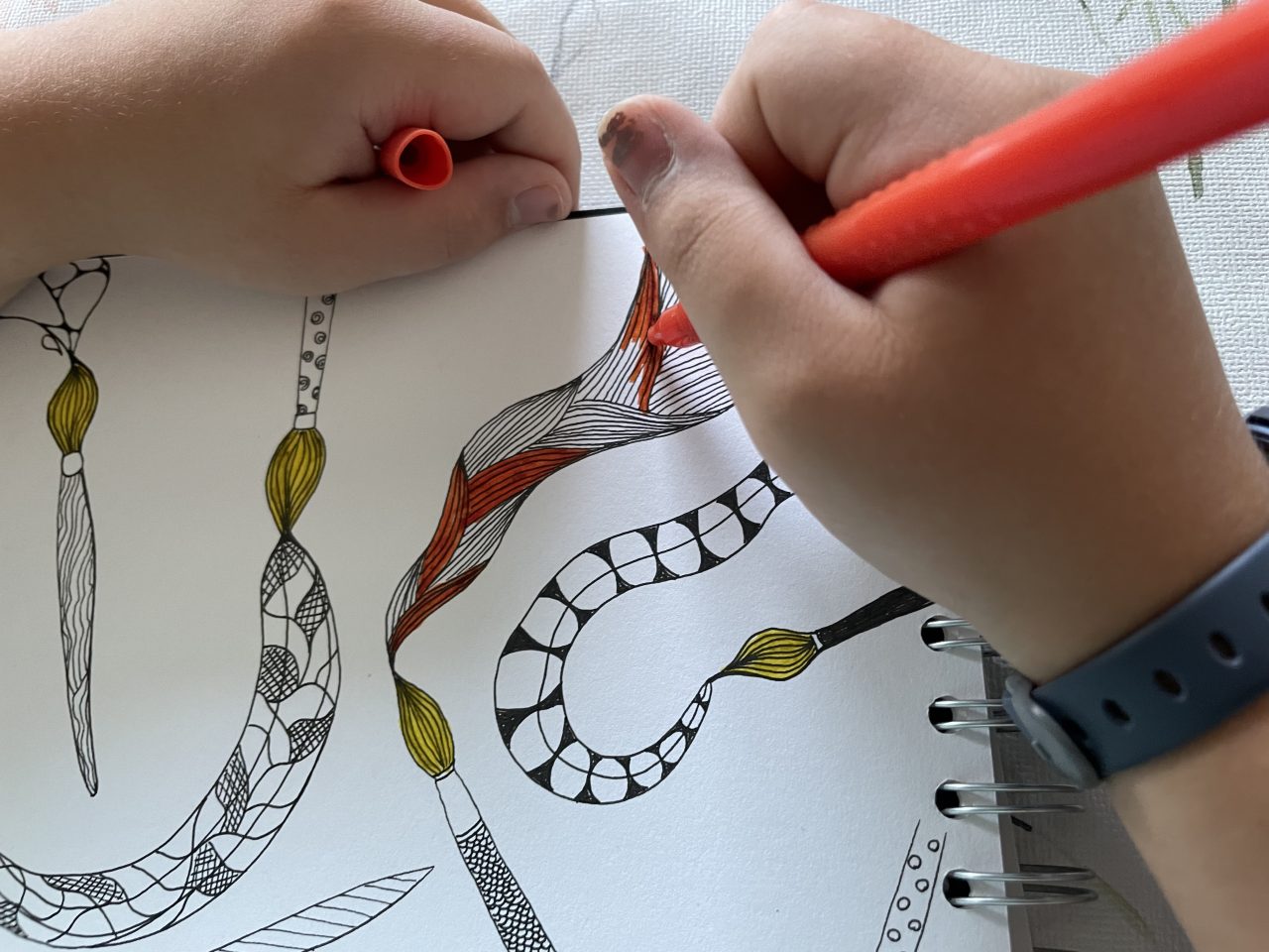 Skizzenblock mit Doodlestiften und Mustern, die bunt ausgemalt werden. Eine Kinderhand hält einen roten Stift und die andere die Kappe. 
