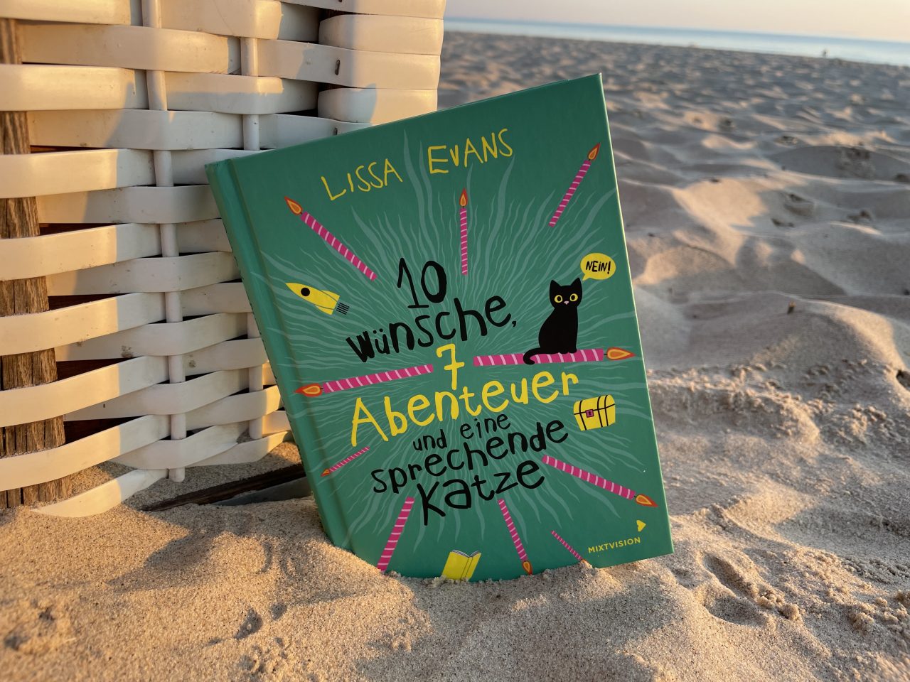 das Kinderbuch "10 Wünsche, 7 Abenteuer und eine sprechende Katze" an einen Strandkorb gelehnt im Sand mit dem Meer im Hintergrund