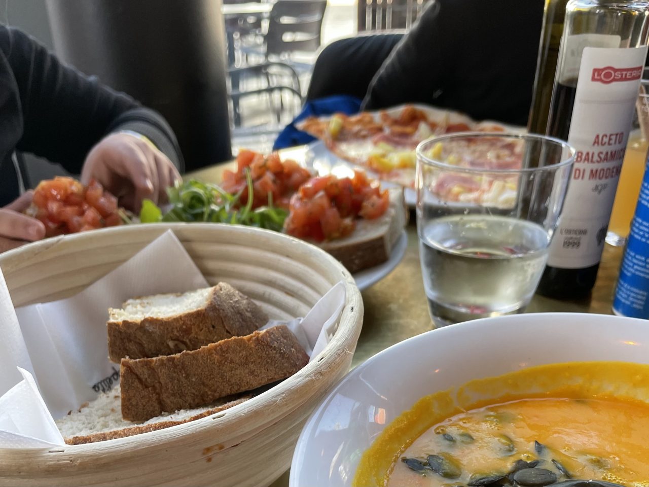 Kürbissuppe, Brot im Korb, Tomatenbrot, Pizza mit Getränken und Ölflaschen auf einem Tisch und im Hintergrund sind teilweise zwei Kinder zu erkennen