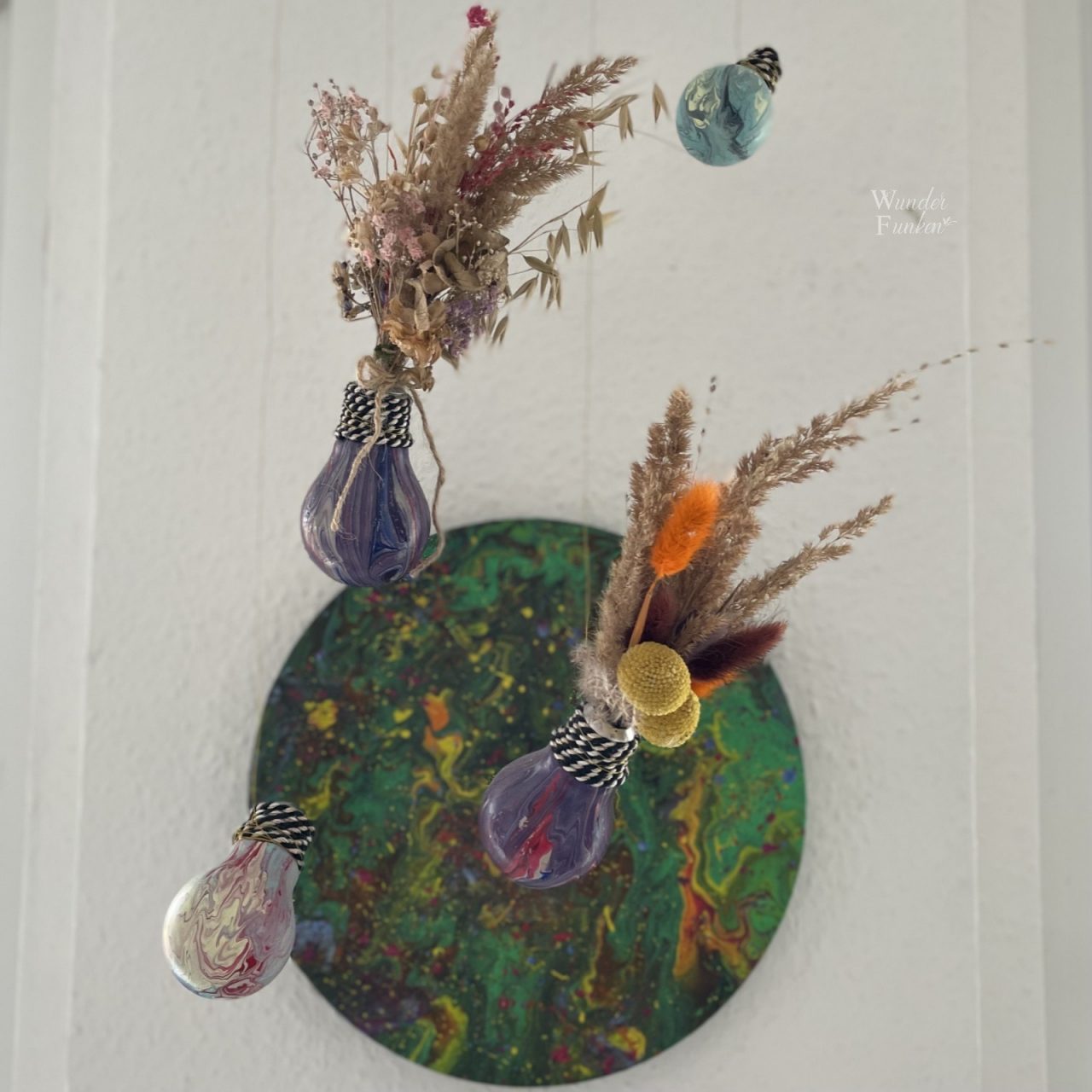 Vier hängende Glühbirnen mit Fluidart gestaltet, zwei davon mit Trockenblumen und ein rundes Fluidartbild an der Wand