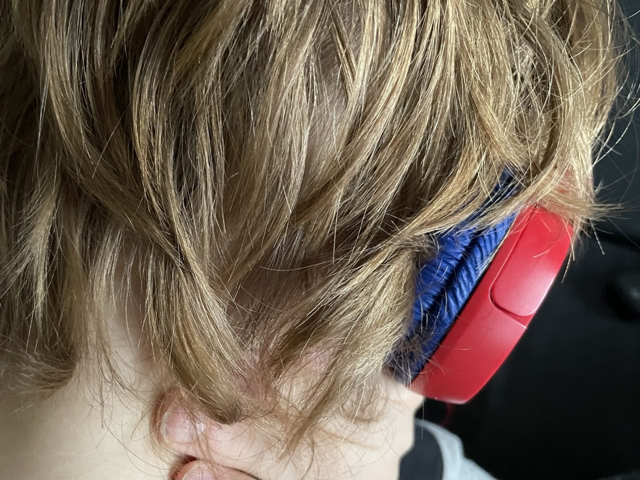 Hinterkopf eines Kindes mit kürzeren blonden Haaren und Hals; dazu ein roter Kopfhörer