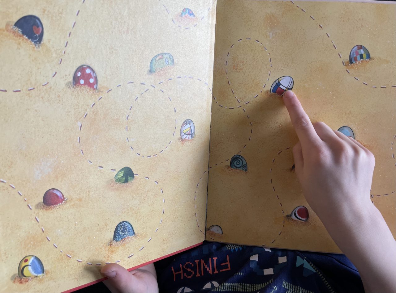 bunte Ostereier im Sand verteilt liegend auf der Seite eines Kinderbuches  und ein FInger zeigt auf eines