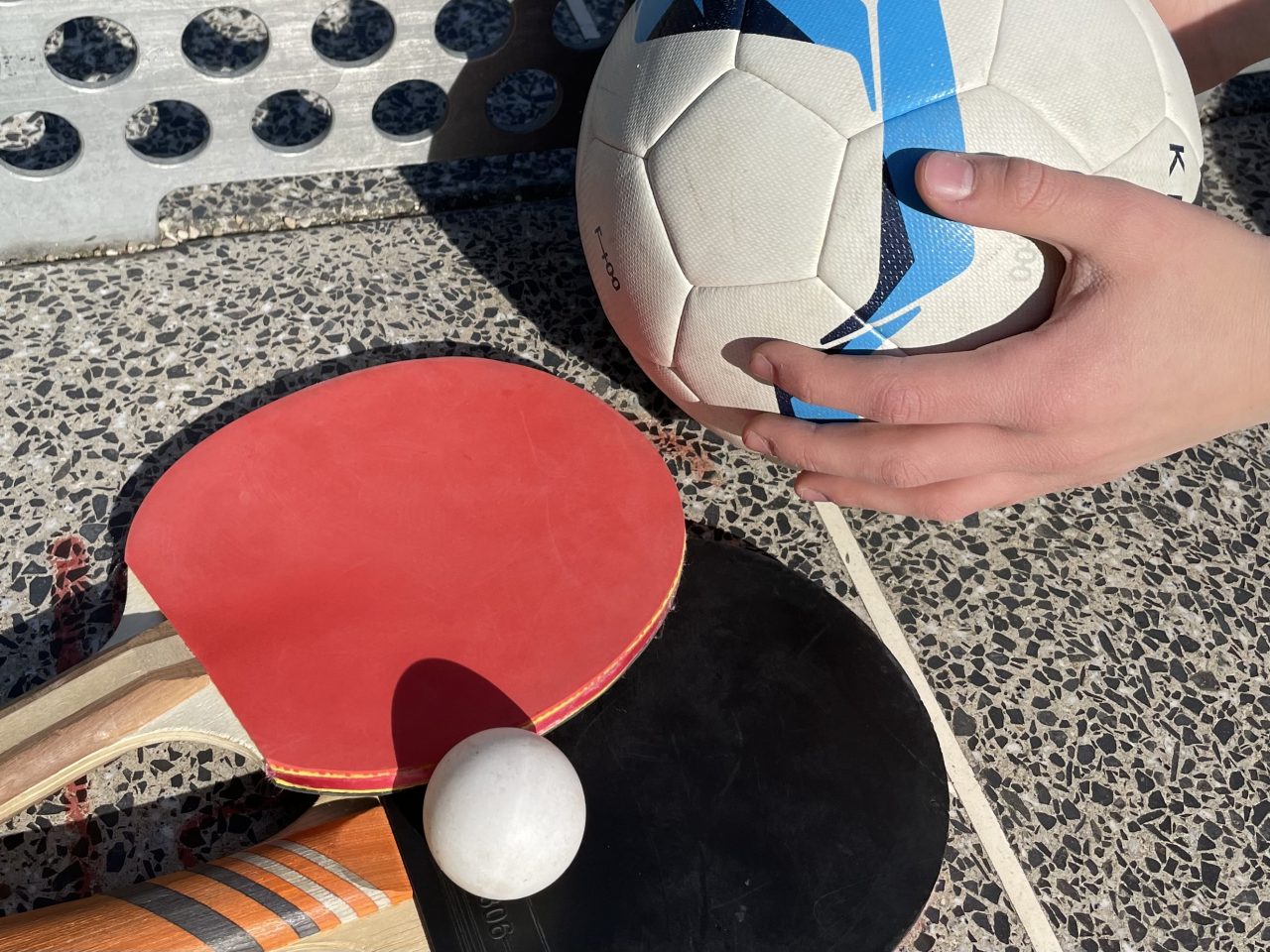 zwei Tischtenniskellen mit einem weißen tischtennisball auf einer Tischtennisplatte liegend - daneben halten Kinderhände einen Fußball