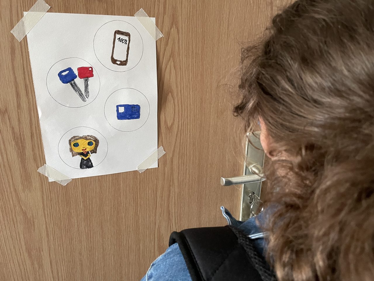 an einer Wohnungstür klebt ein A4-Zettel mit Kreisen, die ein Handy, Schlüssel, eine Fahrkarte und ein Frau zeigen, davor steht ein Mädchen mit Rucksack und Jacke
