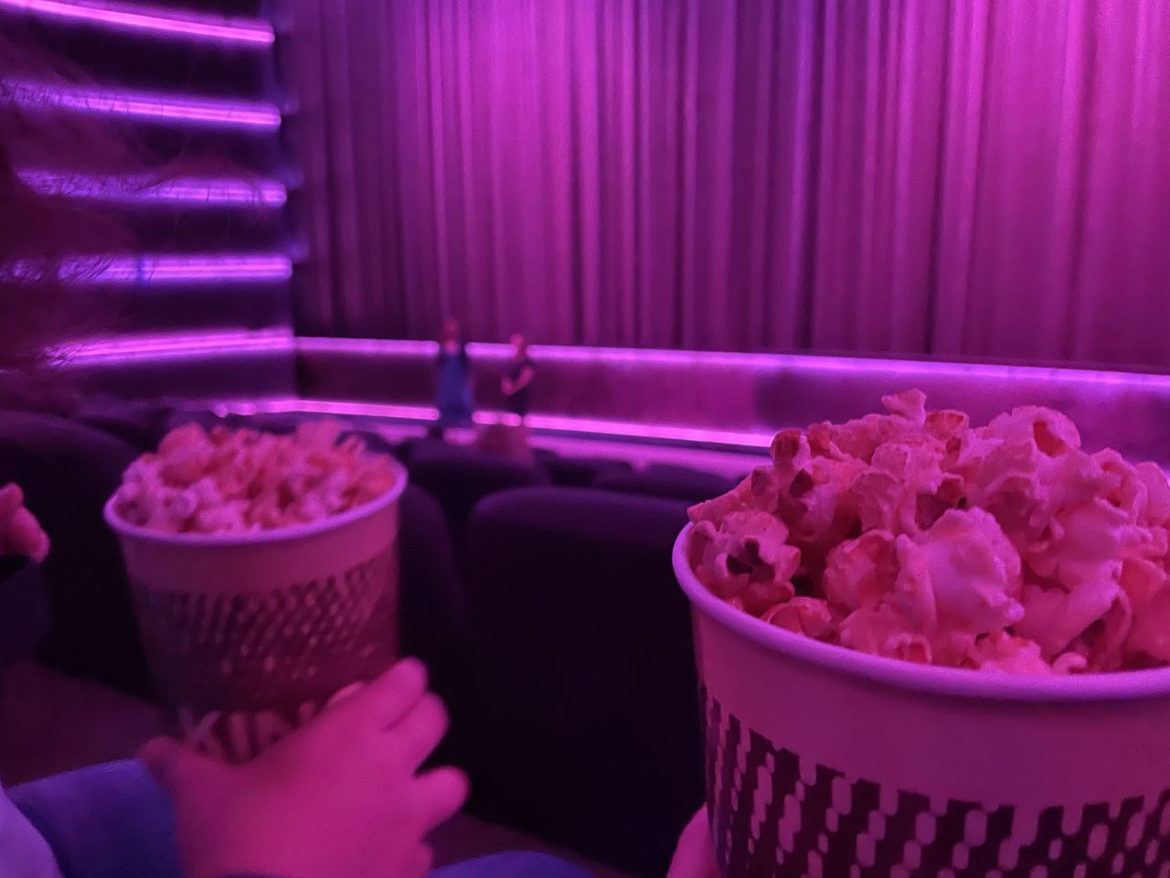 ein in rosa getauchter Kinosaal mit Vorhang vor der Leinwand; zwei gefüllte Popcornbecher im Vordergrund