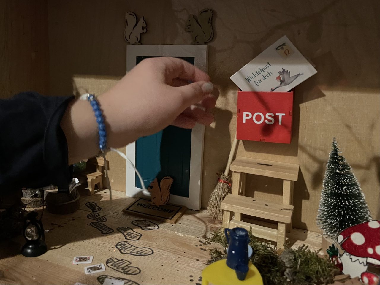in einer Holzkiste ist eine Wichteltür mit roten Briefkasten angebracht und eine Kinderhand greift nach der Wichtelpost