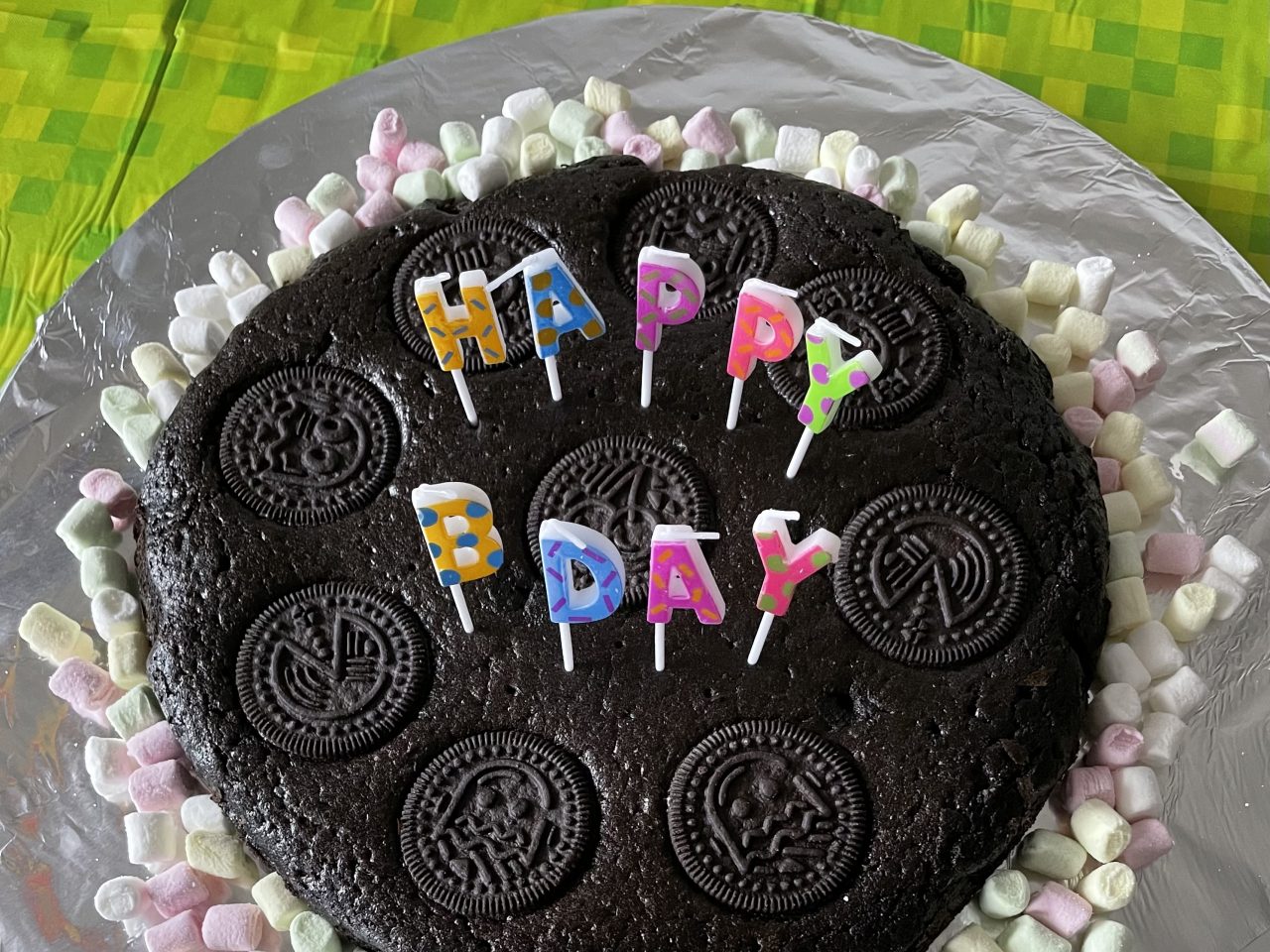 Oreo-Geburtstagskuchen mit "Happy Bday"-Kerzen und umgebenvon  Minimarshmallows auf einem silbernen Teller und grüner Tischdecke