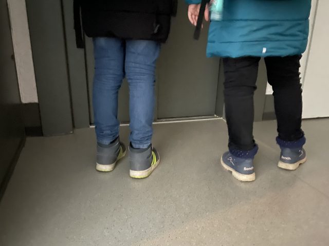 zwei Kinder mit Winterjacken stehen vor einem Fahrstuhl - sie sind von hinten und zur Hälfte zu sehen