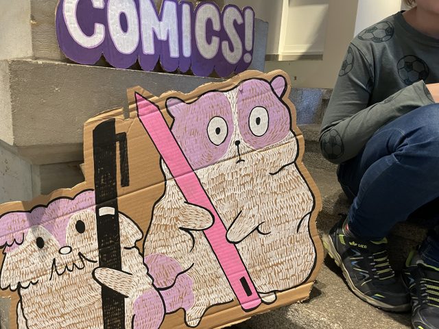 Comic-Hamster gemalt auf Pappe sowie Comic auf Pappe stehend und daneben auf der Treppe ein Kind