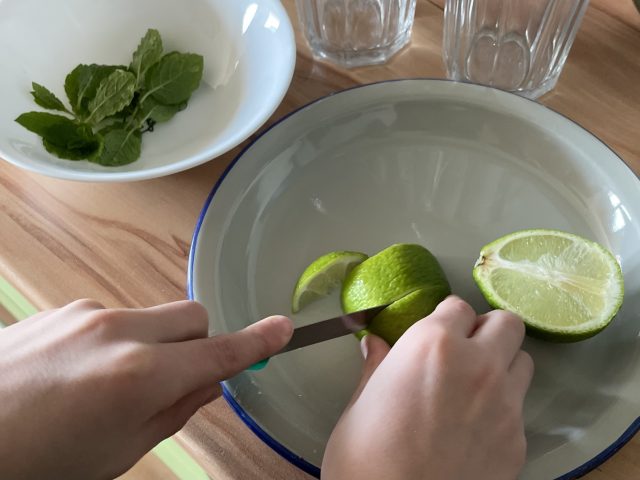 Kind schneidet Limetten auf einem Teller neben einer Schüssel mit Minze