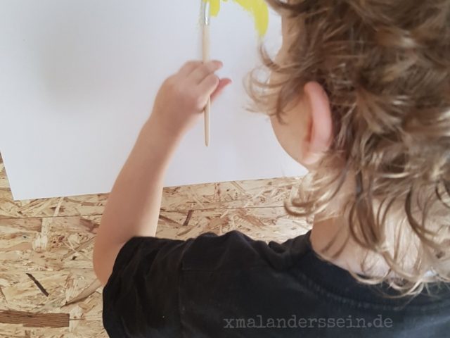 Kind hält einen Pinsel mit gelber Farbe auf einem Papier an der Wand